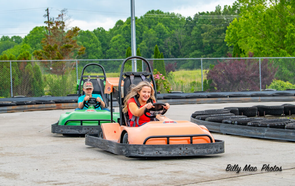 A boy in a green go-kart and a girl in an orange go-kart race around a go-kart track.